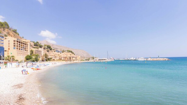 A beach with clear blue water, a sandy shore, and buildings along the coastline. - - BayAltea (Altea)