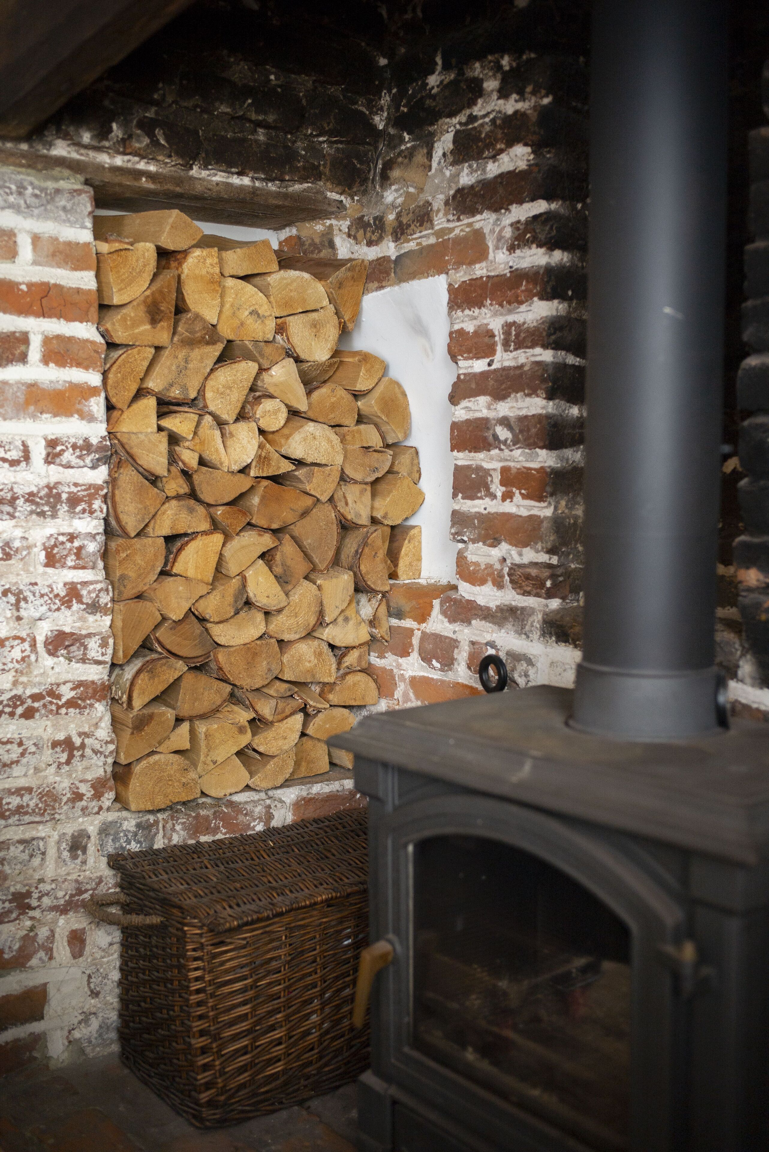 A rustic room with a brick wall, a stack of firewood, a wicker basket, and a wood-burning stove.