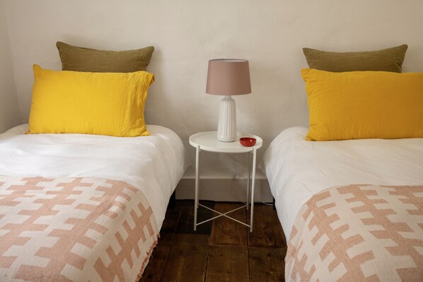 A room with two beds, each with a patterned bedspread and pillows, a small white table with a lamp, and a red object on the table. - Farrier Beach Cottage (Deal)