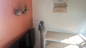A room with a wooden table, a hanging plant, a vacuum cleaner, and a wall-mounted sign with words.