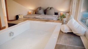 A modern bathroom with a large bathtub, a bedside table with a plant, and a window with a view.