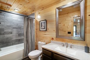 A bathroom with a wooden ceiling, a large mirror, a white bathtub, a toilet, and a sink with two faucets.