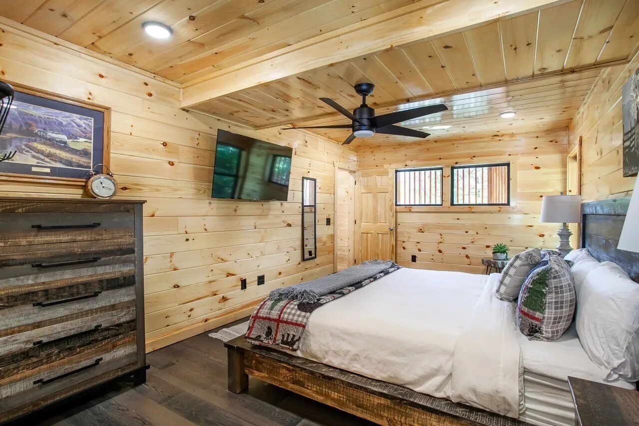 A wooden cabin bedroom with a bed, a ceiling fan, a mounted TV, and a wooden dresser.