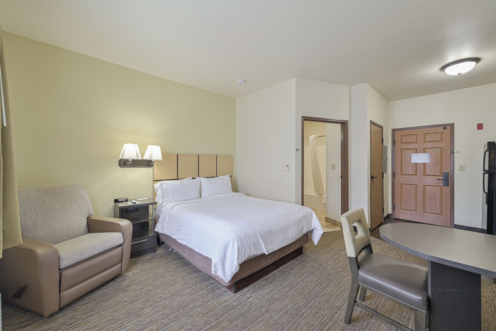 Habitación de hotel con cama, escritorio, silla, mesita de noche, lámpara, cortina de ducha y puerta.