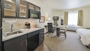 Ein Hotelzimmer mit Küchenzeile, einem Bett, einem Schreibtisch, einem Stuhl, einem Fernseher und einem fenster mit Vorhängen.