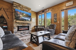 A cozy living room with a stone fireplace, leather sofas, a television, and a large window offering a scenic view.