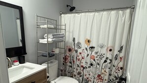 Et badeværelse med en hvid vask, toilet, bruseforhæng med blomstermønster, en metalkasse med håndklæder og et spejl.