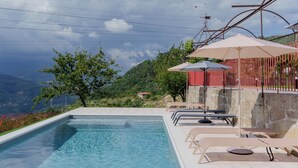 Une piscine entourée de chaises longues et d’un mur de pierre, ainsi qu’une clôture rouge, avec vue sur des collines et un ciel nuageux.