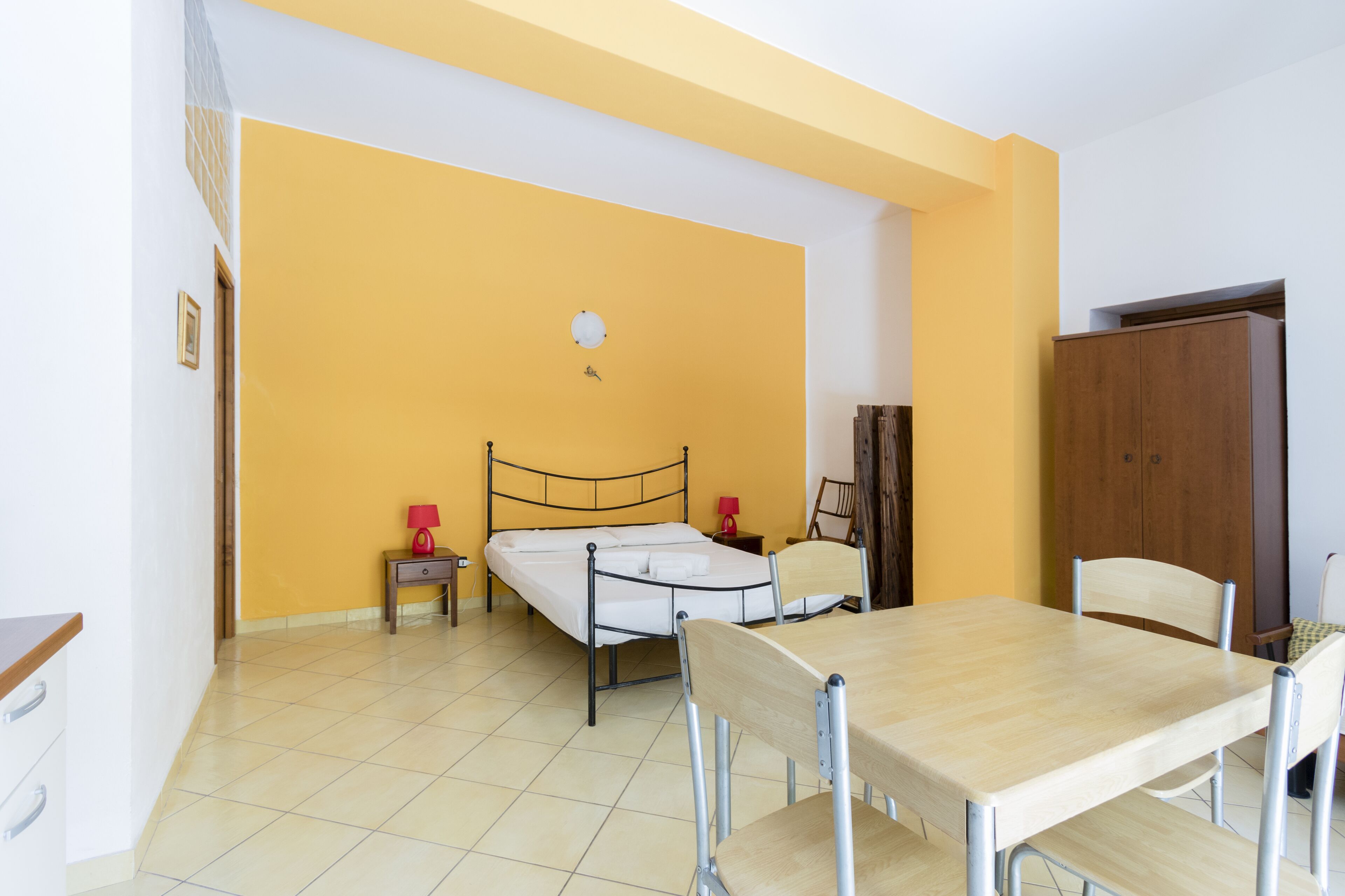 A room with a bed, a wooden table, and a chair. There is a wardrobe and a small table with a lamp.