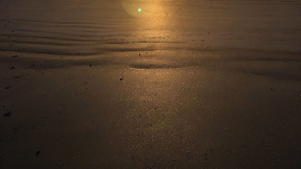 Una playa con un atardecer sobre el océano, un barco a lo lejos y huellas en la arena.
