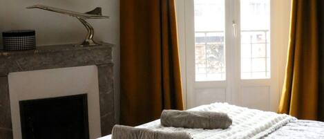 Un dormitorio con cama, chimenea y vistas a través de una ventana con cortinas.