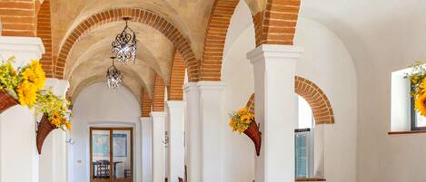 Un pasillo con techos altos de ladrillo abovedado, paredes blancas y candelabros colgantes. A ambos lados del pasillo hay cesteras de mimbre con flores.