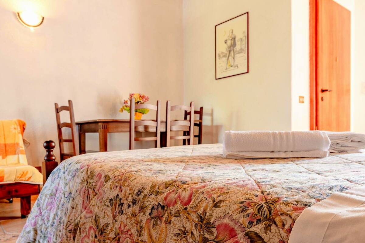 Ein Schlafzimmer mit geblümter Bettdecke, einem Holzstuhl, einem kleinen Tisch mit Blumen und einem gerahmten Bild an der Wand.