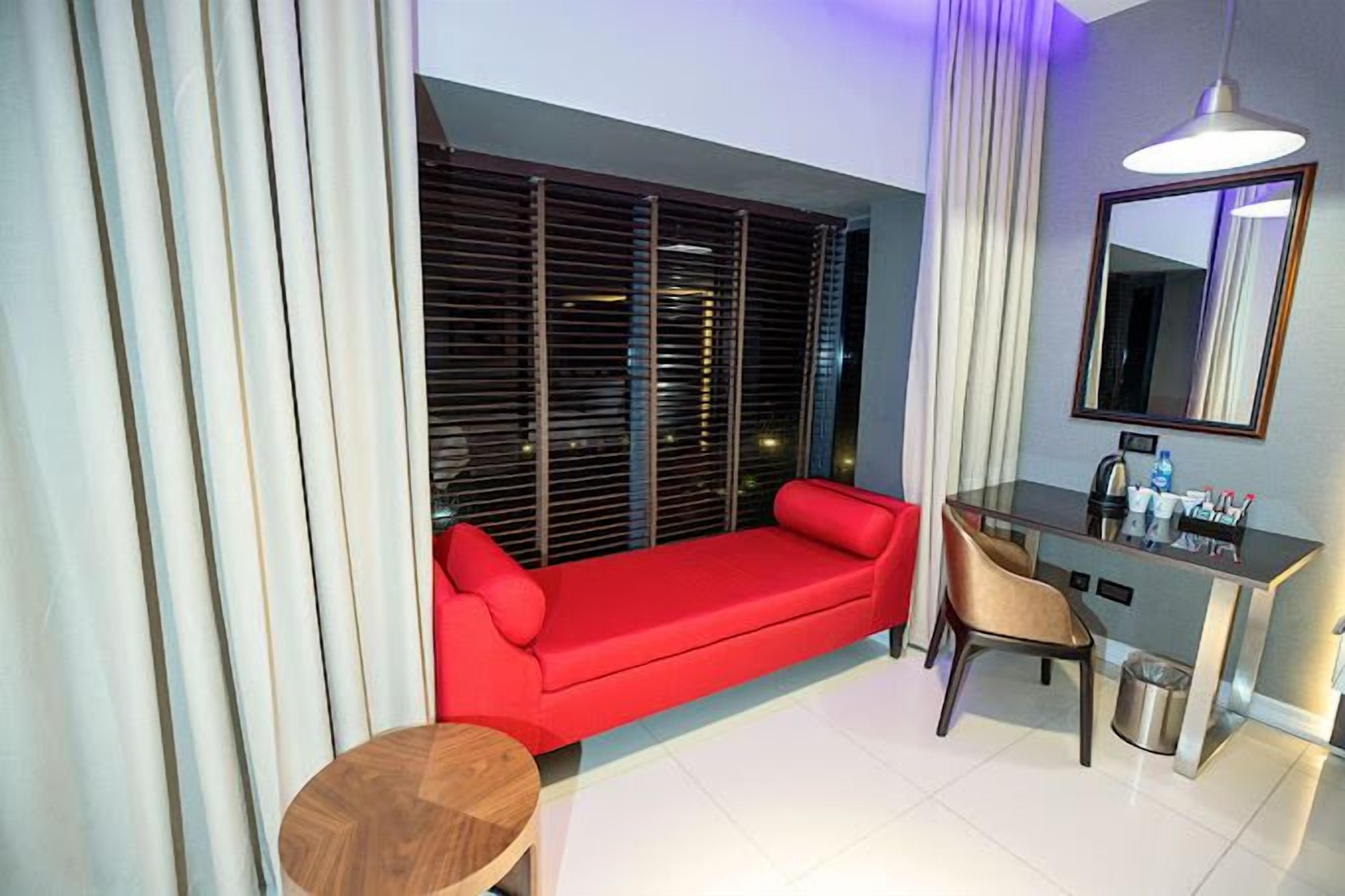 Ein modernes Hotelzimmer mit einem roten Sofa, einem kleinen runden Tisch, einem Schreibtisch mit Stuhl und einem großen Fenster mit Jalousien.