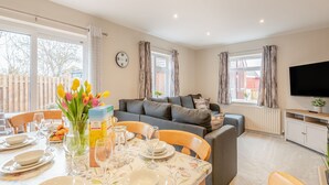 A well-lit living room with a dining area, a grey sofa, a flat-screen TV, and a clock on the wall.