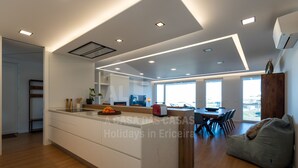 A modern kitchen with a central island, a dining area with a wooden table and chairs, and large windows providing natural light. - Casa Mango by Acasadascasas (Mafra)