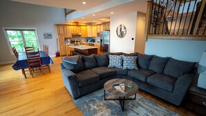 A modern living room with a sectional sofa, a dining table with chairs, and a kitchen in the background. - #5 Lakefront Hamlin Lake Retreat - Sleeps 10 (Ludington)