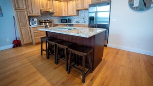 A modern kitchen with wooden cabinets, a stainless steel refrigerator, a granite island with bar stools, and a wall clock. - #5 Lakefront Hamlin Lake Retreat - Sleeps 10 (Ludington)