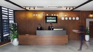 Uma área de recepção com um balcão de madeira, dois recepcionistas, uma televisão e uma fileira de relógios na parede.