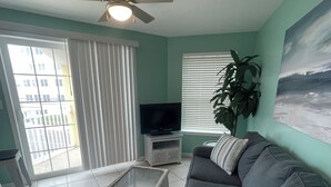 A living room with a sofa, a television, a ceiling fan, and a window with blinds.