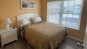 A bedroom with a bed, a nightstand, a lamp, a painting on the wall, and a window with blinds.