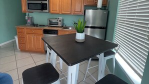 A compact kitchen with wooden cabinets, a microwave, a toaster, a refrigerator, and a dining area with white stools.
