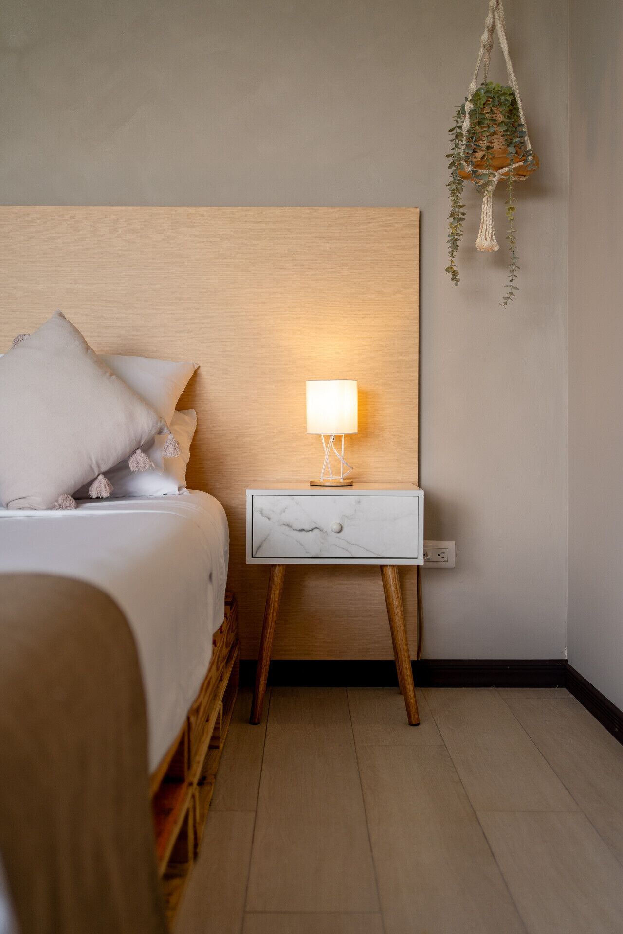 Un dormitorio ordenado con cama, mesita de noche y una planta colgante.