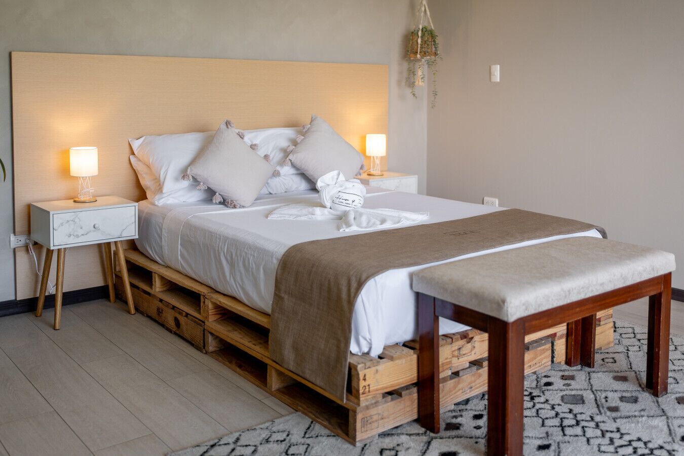 Una cama con cabecera de madera, dos mesitas de noche con lámparas y un banco al pie de la cama.