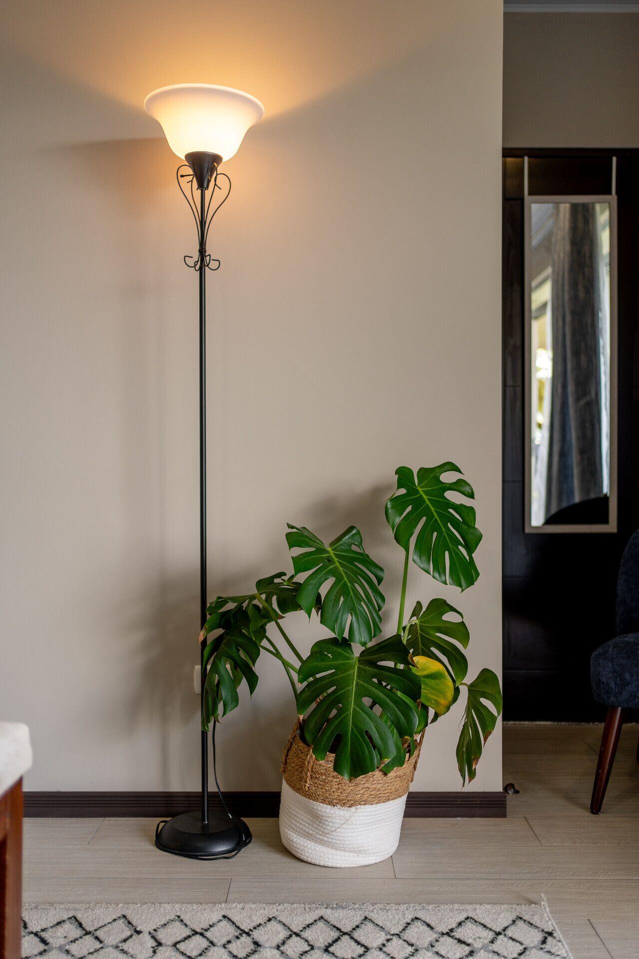 Een vloerlamp, een pot met monstera en een stoel in een kamer met een kamerbreed tapijt.