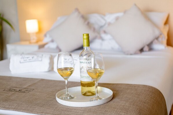 A bed with white linens and pillows, a bedside table with a bottle and glasses of white wine, and a nightstand with a lamp.