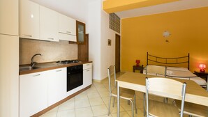 A compact kitchen with white cabinets, a stove, and a sink. There is a dining table with chairs and a bed with a headboard in the adjacent room. - Visitsicilia Le Magnolie 2 (Castellammare del Golfo)
