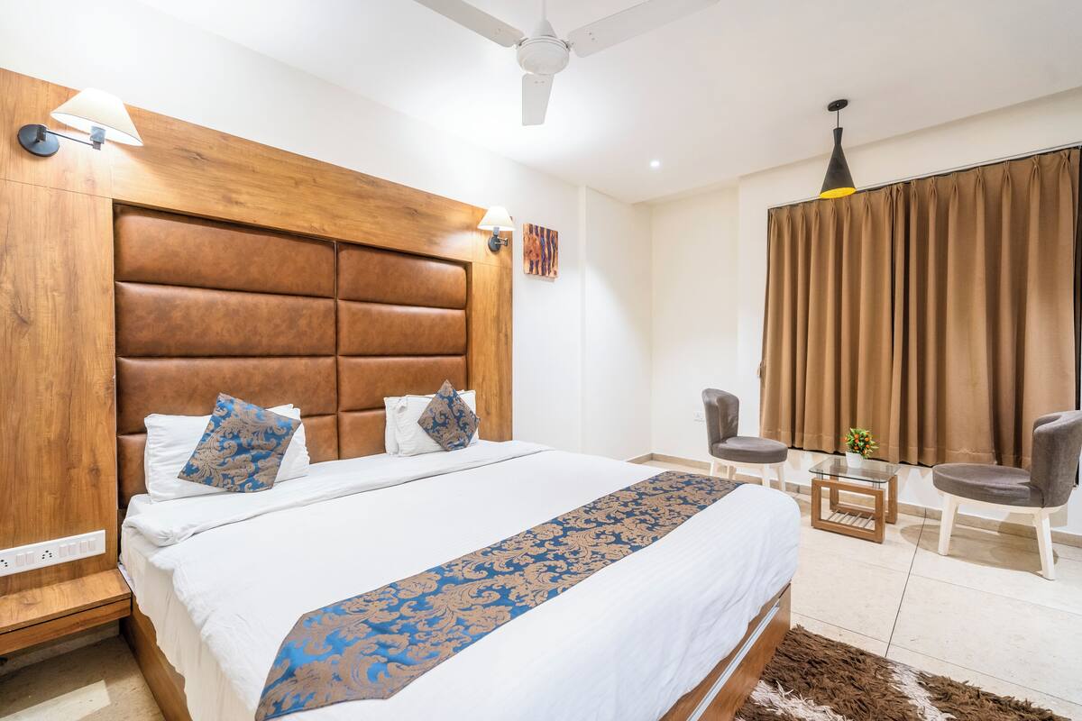 A hotel room with a large bed, a wooden headboard, a bedside table, a chair, a small table with a plant, and a ceiling fan.