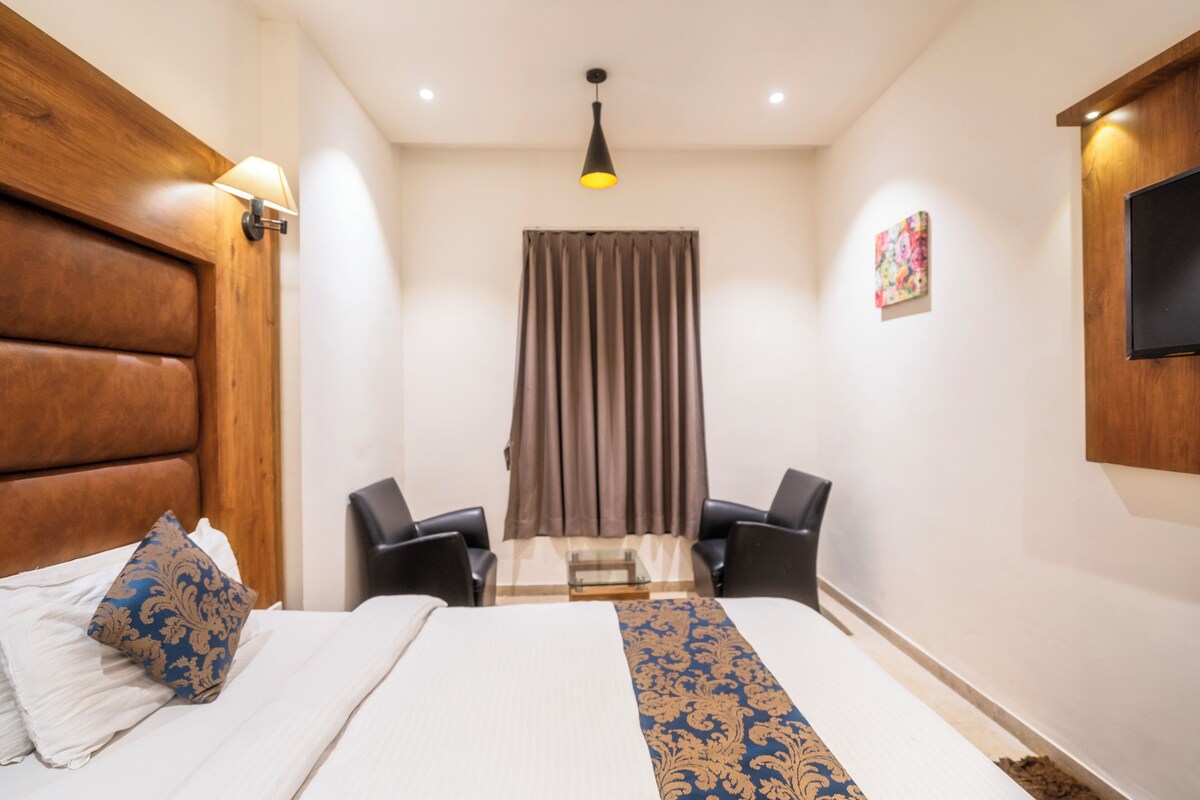 A hotel room with a large bed, two chairs, a television, a window with curtains, and a wooden headboard.