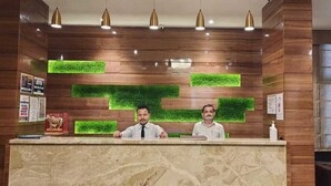 A reception desk with two staff members, wooden paneling, green vertical elements, and a window with blinds. - Hotel Bhabha (Rajkot)