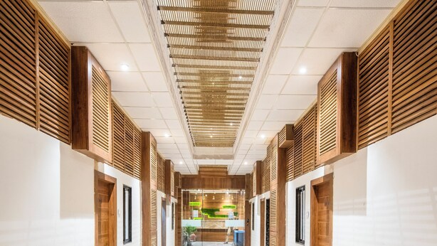 A corridor with a tiled floor, wooden paneling on the walls, and a ceiling with a grid pattern. - Hotel Bhabha (Rajkot)
