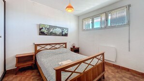 A bedroom with a wooden bed, a nightstand, a wall-mounted lamp, a window with lace curtains, and a painting of orchids.