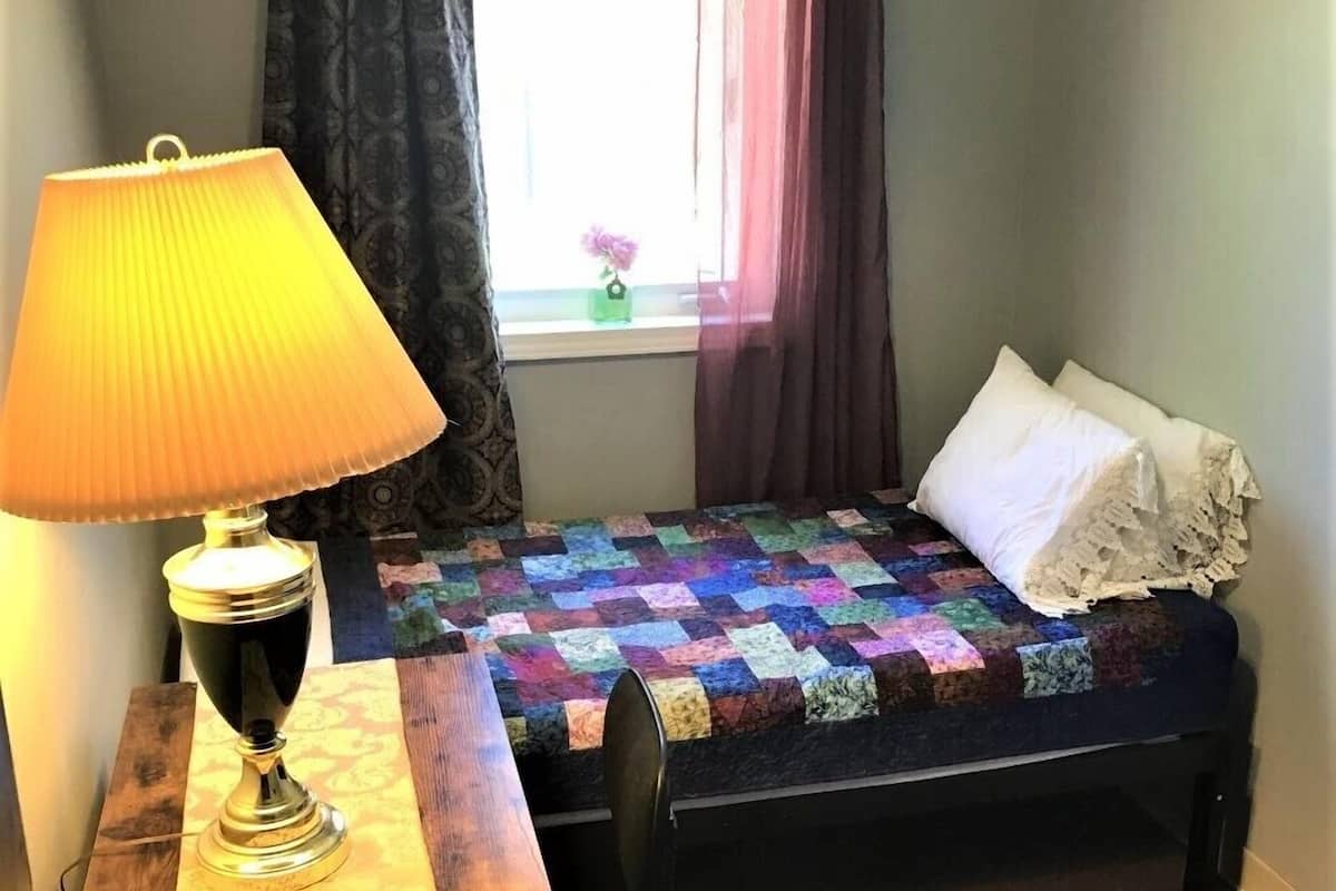 A small bedroom with a bed, a desk with a lamp, a window with curtains, and a quilt on the bed.