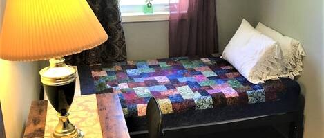 Un dormitorio pequeño con cama, un escritorio con lámpara, una ventana con cortinas y una colcha en la cama.