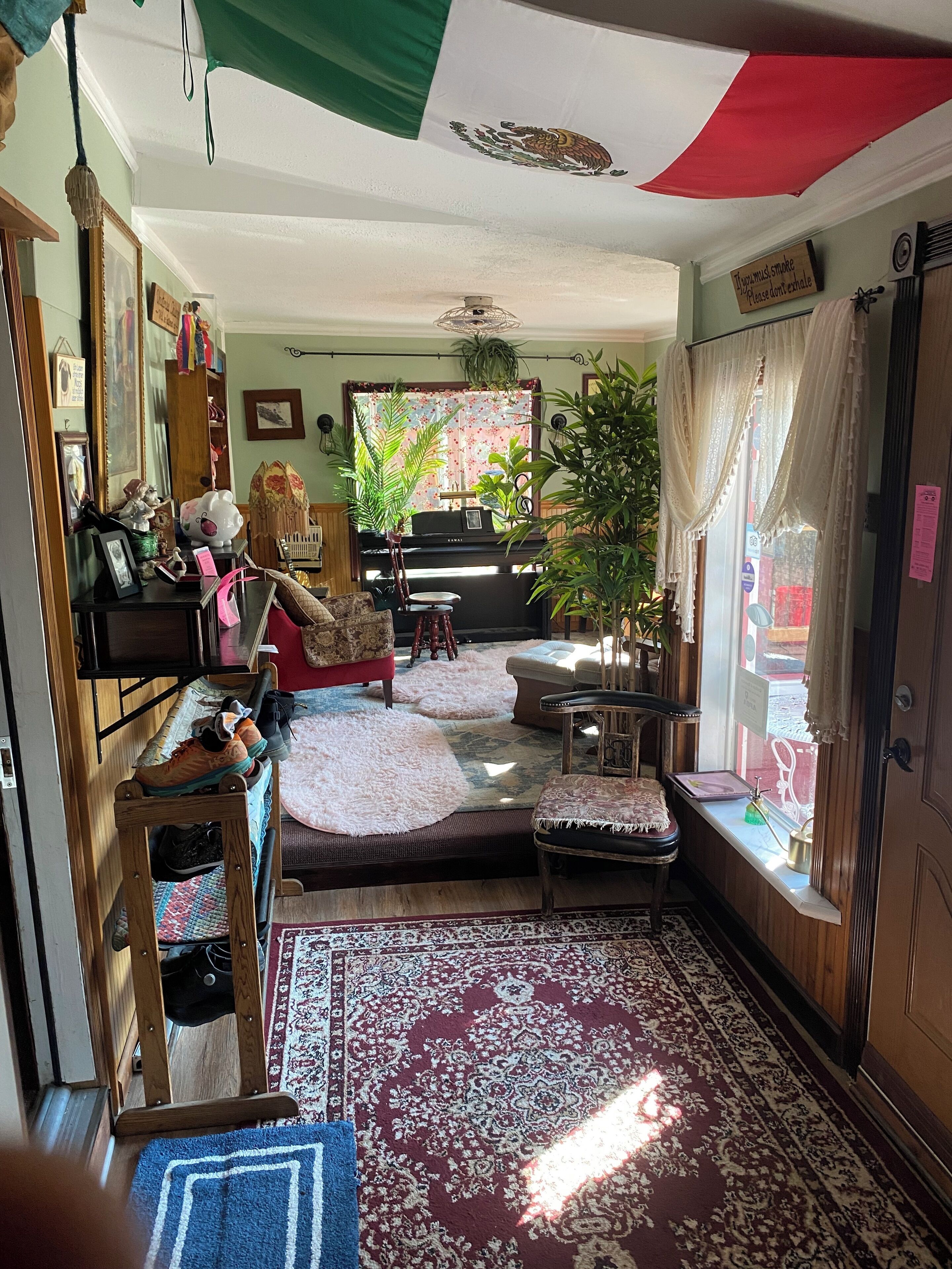 A cozy living room with a piano, a sofa, a chair, and a small table. There is a window with curtains and a plant on the windowsill.