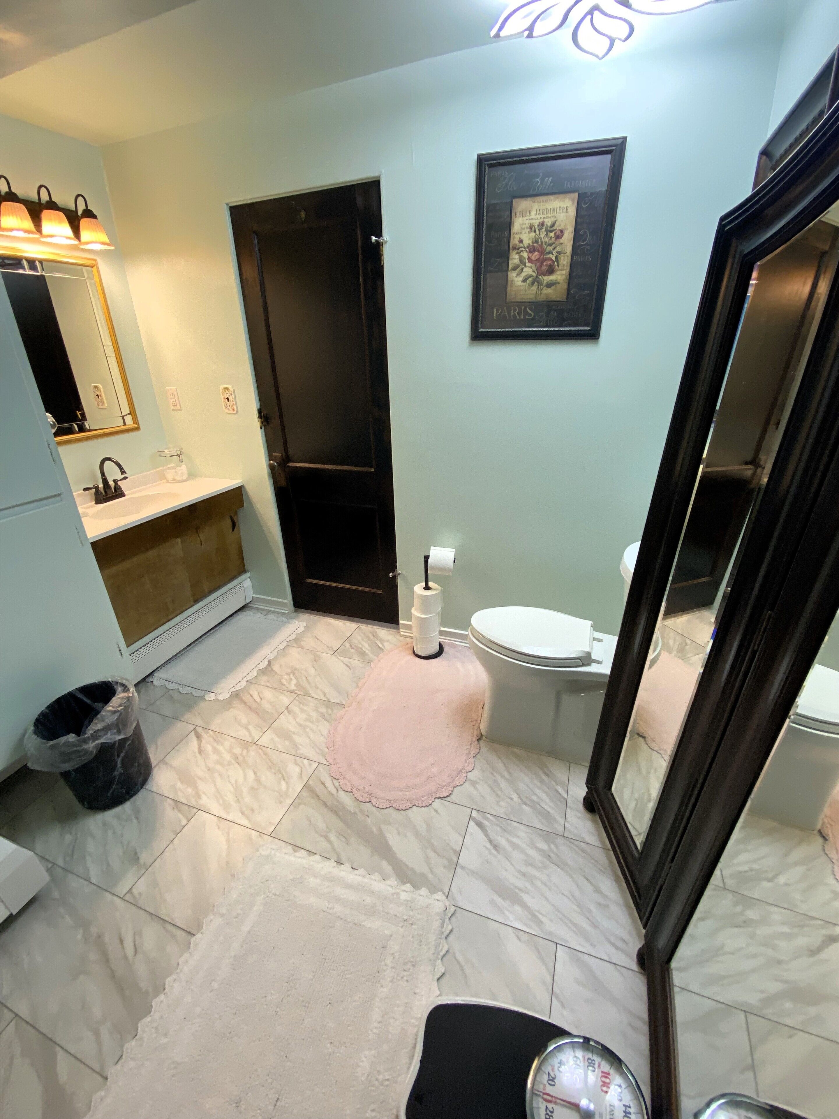 Un baño con lavabo, inodoro y espejo. Hay un cuadro en la pared y un cubo de basura.