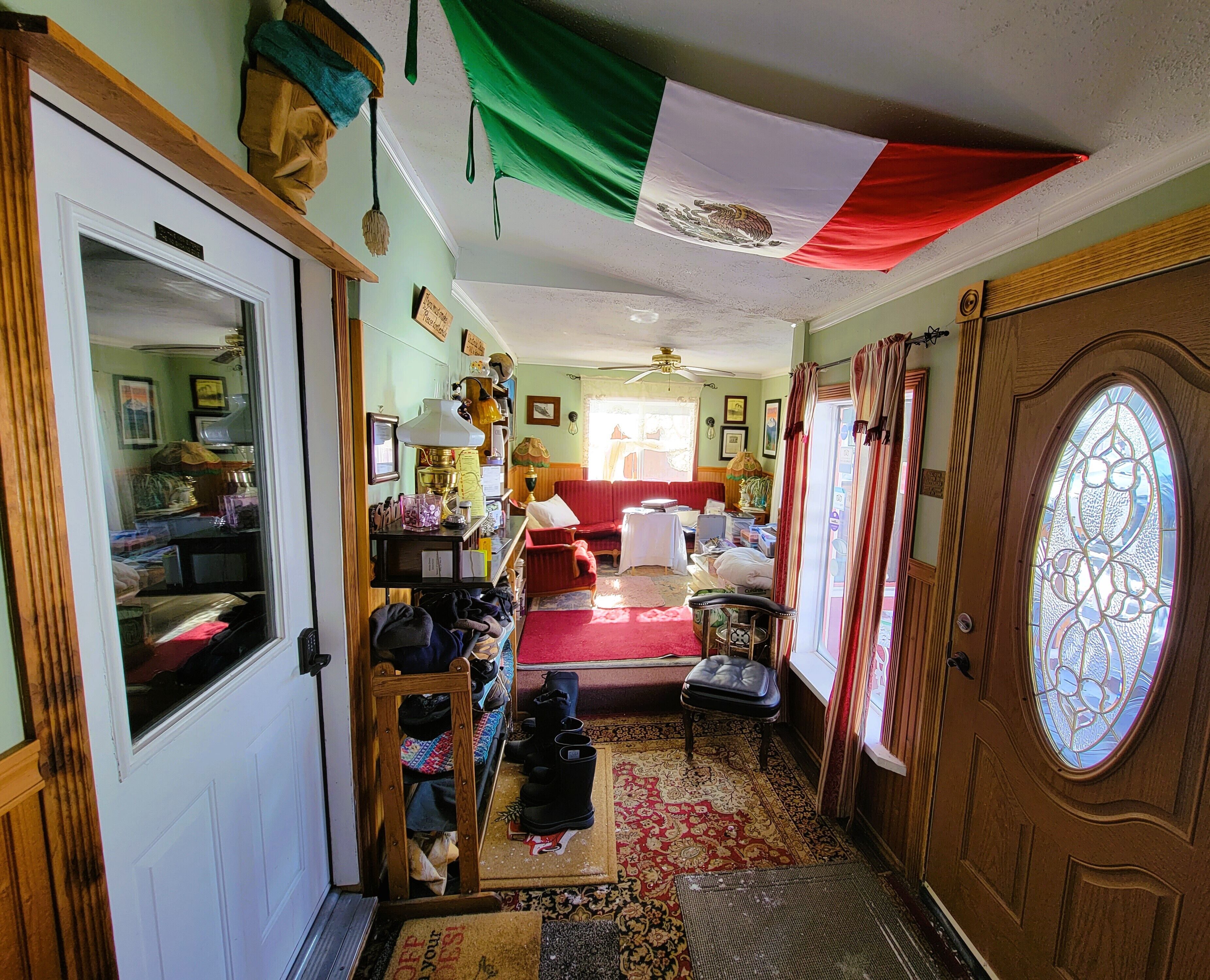 A cozy living room with a red sofa, a wooden chair, and a patterned rug. A wooden door with a glass panel is visible, and a flag hangs from the ceiling.