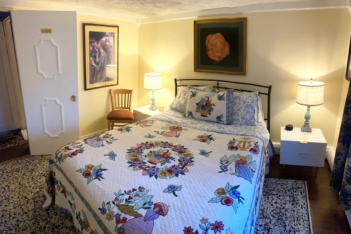 A bedroom with a floral bedspread, two bedside lamps, a framed picture of a rose, and a chair.