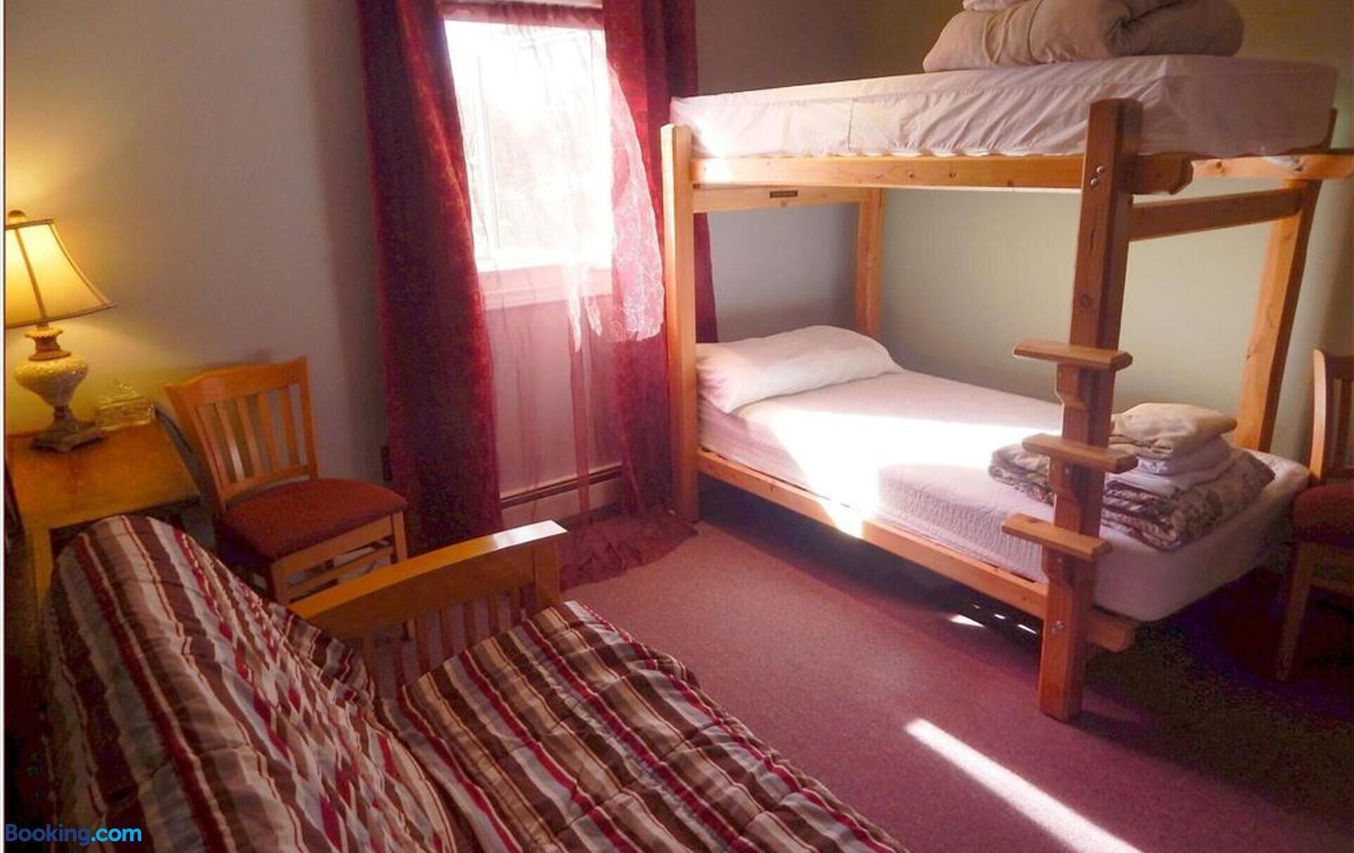 A bunk bed room with a single bed, a chair, a table with a lamp, and a window with curtains.
