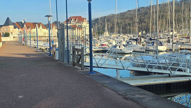 A marina with numerous boats docked, a paved walkway, and buildings in the background. - Nice house with garden near the beach (Dives-sur-Mer)