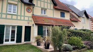 A traditional half-timbered house with a sloped roof, a front entrance with green shutters, and a small garden with various plants. - Nice house with garden near the beach (Dives-sur-Mer)