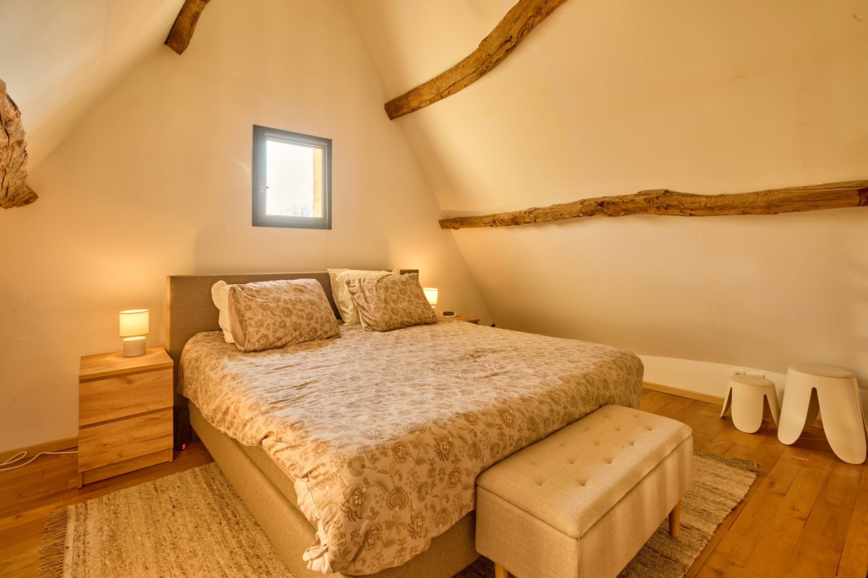 A bedroom with a large bed, wooden headboard, bedside tables with lamps, a window, and a sloped ceiling with exposed beams.