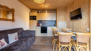 A cozy living area with a dark brown sofa, wicker chairs, and a dining table with a TV mounted on the wall.