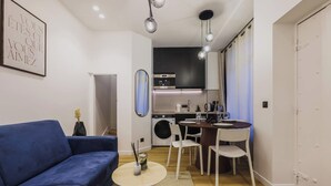 Een knus woongedeelte met een blauwe bank, een klein rond tafeltje, een keukenhoek met een wasmachine, en een eettafel met stoelen.