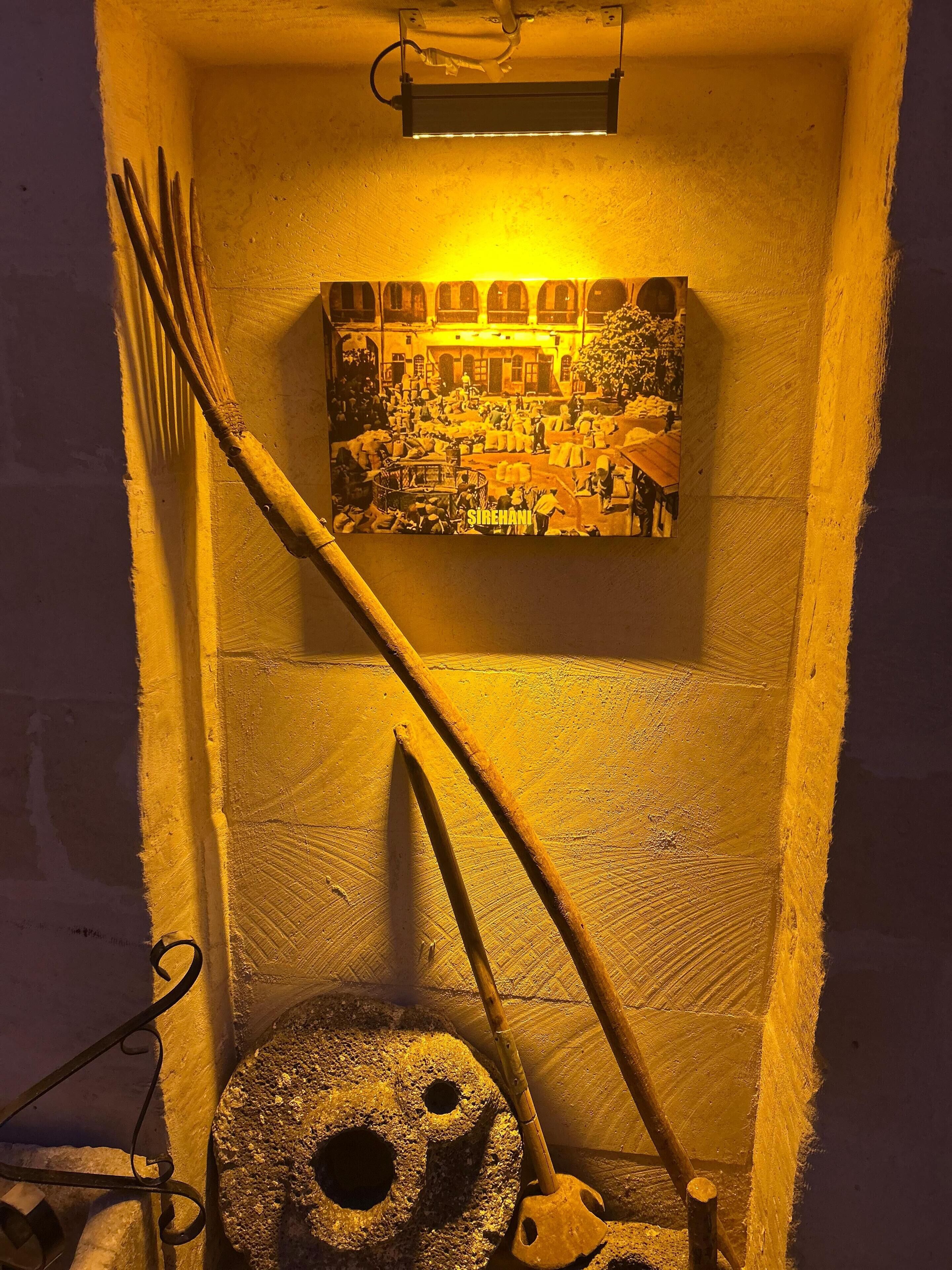 Una habitación con decoración rústica, que presenta un rastrillo de madera apoyado en la pared y una piedra giratoria.