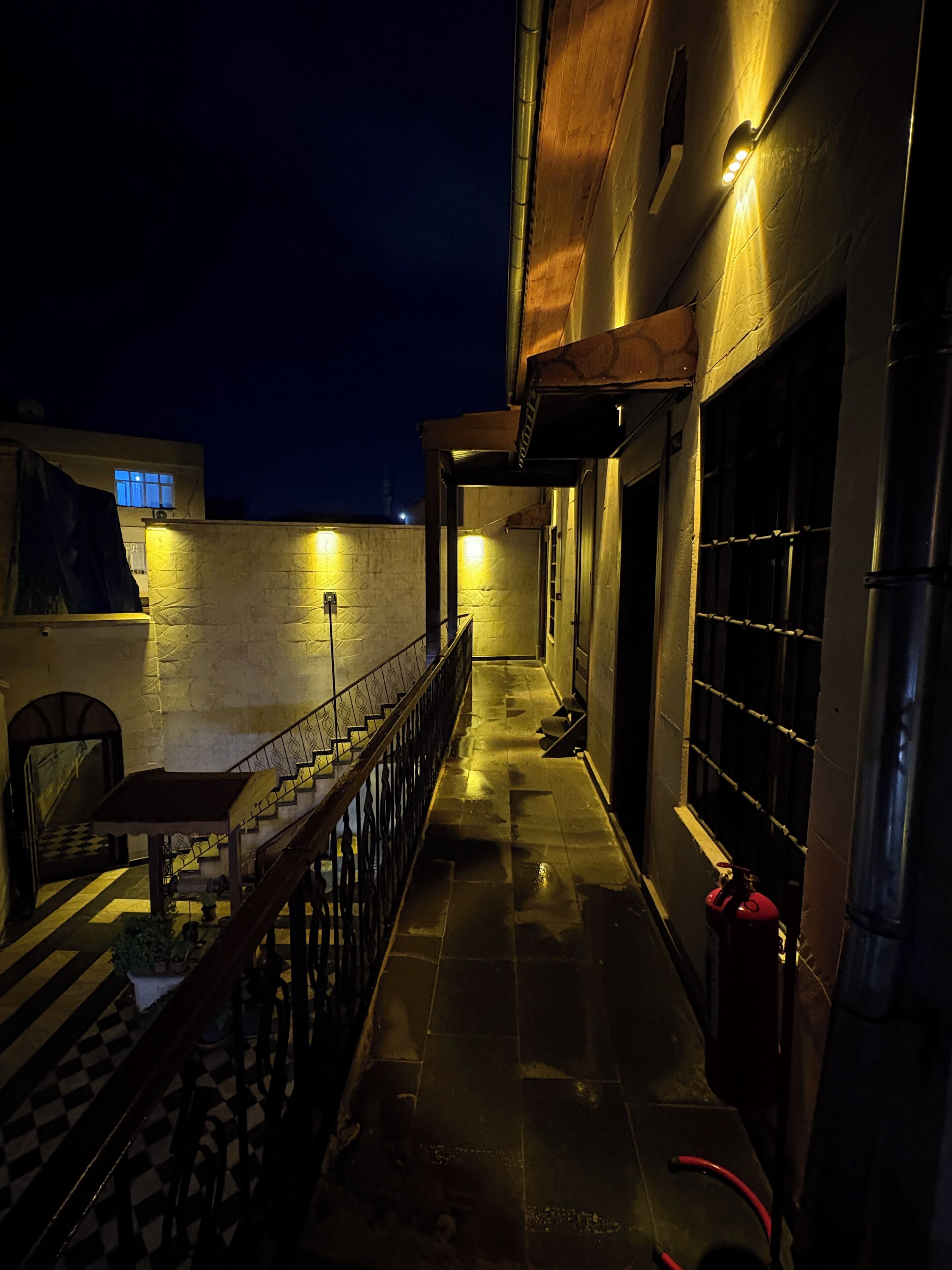 A narrow, dimly lit corridor with a checkered floor, a metal railing, and a fire extinguisher.